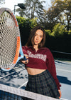Alisha Marie wearing a cropped Maroon Vlogmas 2021 Quarter Zip Sweatshirt. Screen print front and sleeve in color cream. She is wearing a skirt and has a tennis racket.